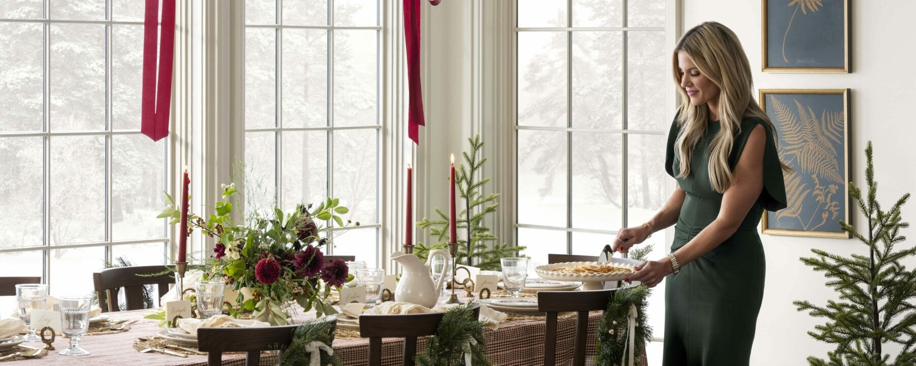 A woman in a dark green dress sets a pie on a festive dining table decorated with greenery, flowers, and holiday accents near large windows on a snowy day. Small Christmas trees and red ribbons add to the cozy holiday atmosphere, by McGee & Co.