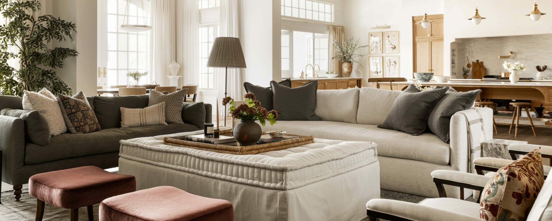 A spacious, sunlit living room with large windows, neutral-tone sofas, a padded ottoman coffee table, accent pillows, two pink stools, and a view into an open kitchen and dining area in the background, by McGee & Co.